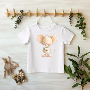 Może przedstawiać: Biała koszulka z kreskówkową myszą trzymającą stokrotkę. Mysz ma duże uszy i puszysty korpus, nosi różowe buty. Koszulka wisi na drewnianym stojaku z zielenią.