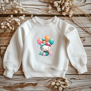 Könnte beinhalten: Weißes Sweatshirt mit einem Cartoon-Katzen-Design, das Luftballons hält. Die Katze trägt einen Partyhut und ist von Konfetti umgeben. Das Sweatshirt hat lange Ärmel und einen Rundhalsausschnitt.