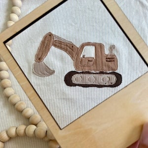 Puede incluir: Aplicación textil enmarcada que representa una excavadora marrón sobre un fondo crema acanalado. La excavadora tiene un diseño detallado con un brazo, una pala y orugas marrones. El marco es de madera clara.