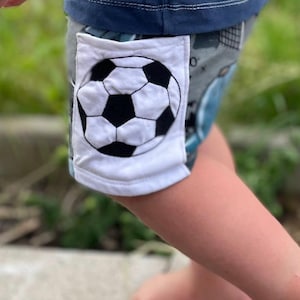 Könnte beinhalten: Shorts mit einem dunkelblauen Bund und einem grau gemusterten Hauptstoff. Ein weißer, quadratischer Aufnäher ist an der Seite angebracht und zeigt eine schwarz-weiße Fußballgrafik. Das Kleidungsstück weist sichtbare Nähte am Bund und am Aufnäher auf.