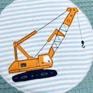 May include: Embroidered patch featuring a yellow construction crane on a light blue and white striped background. The crane has a black hook and is set against a textured, teal-colored fabric. The patch is circular with a white border.