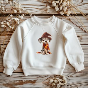 Könnte beinhalten: Weißes Sweatshirt mit einem Cartoon-Dalmatiner-Welpen, der einen braunen Zaubererhut und einen rot-goldenen Schal trägt. Der Welpe hat schwarze Flecken und sitzt. Das Sweatshirt hat lange Ärmel und einen Rundhalsausschnitt.