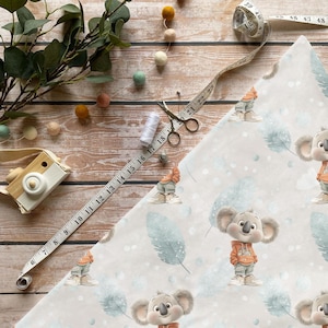 Könnte beinhalten: Hellfarbener Stoff mit Koala-Bären-Muster, Maßband, Schere und Faden. Der Stoff zeigt Koala-Illustrationen mit orangefarbenen Kapuzenpullovern und grauen Hosen. Eine Spielzeugkamera aus Holz und dekorative Filzkugeln sind ebenfalls vorhanden.