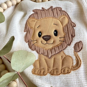 Könnte beinhalten: Ein braun-beiges Löwen-Applikation ist auf einem weißen Waffelstoff zentriert. Der Löwe hat eine braune, gerippte Mähne und einen Schwanz, mit einem beigen Gesicht und Körper. Der Löwe lächelt mit schwarzen Augen und Schnurrhaaren. Ein Eukalyptuszweig und Holzperlen sind im Hintergrund.