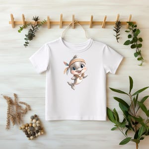 Könnte beinhalten: Weißes T-Shirt mit einem Zeichentrick-Delphin, der ein indianisches Stirnband mit Federn trägt. Der Delphin hat einen freundlichen Ausdruck und ist in einer spielerischen Pose. Das T-Shirt hängt an einem Holzgestell.