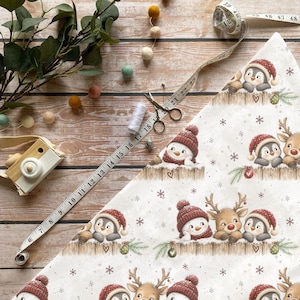 Könnte beinhalten: Ein weißer Stoff mit einem wiederkehrenden Weihnachtsmuster aus Pinguinen, Schneemännern und Rentieren mit roten Mützen, verziert mit Schneeflocken und Ornamenten. Ein weißes Maßband zeigt Maße in cm neben einer Schere.