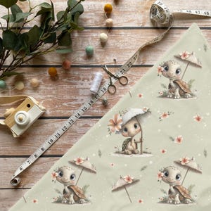 Könnte beinhalten: Hellgrüner Stoff mit einem sich wiederholenden Muster von Zeichentrickschildkröten, die Regenschirme halten und Blumengirlanden tragen. Die Schildkröten sind hellbraun und braun, mit großen Augen. Der Stoff ist mit kleinen Blumen verziert. Ein Maßband ist sichtbar.