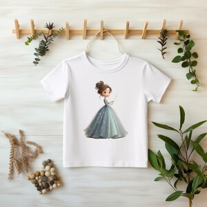 Könnte beinhalten: Weißes T-Shirt mit einer Cartoon-Illustration einer Prinzessin in einem blauen Kleid, die einen gelben Zauberstab hält. Die Prinzessin hat braunes Haar, das zu einem Dutt gebunden ist. Das T-Shirt hängt an einem Holzgestell mit dekorativem Grün.