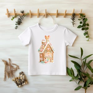 Könnte beinhalten: Weißes T-Shirt mit einer skurrilen Illustration eines Lebkuchenhauses mit bunten Süßigkeiten und einer Maus. Das Haus hat herzförmige Details und einen Schornstein. Ein süßes Design für Kinder.