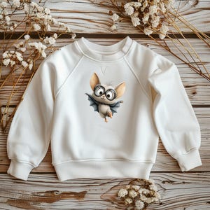 Könnte beinhalten: Weißes Sweatshirt mit einem Cartoon-Fledermaus-Design. Die Fledermaus hat eine große Brille und einen Umhang. Das Sweatshirt hat einen Rundhalsausschnitt und lange Ärmel. Das Design befindet sich mittig auf der Vorderseite.