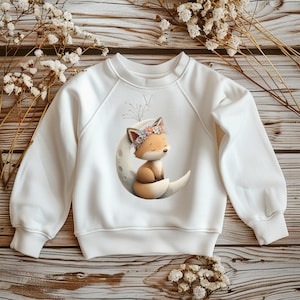 Może przedstawiać: Biała bluza z uroczą ilustracją śpiącego lisa w koronie z kwiatów, siedzącego na księżycu w kształcie półksiężyca. Lis jest w odcieniach brązu i pomarańczy, z miękkim, marzycielskim designem. Bluza ma długie rękawy i okrągły dekolt.