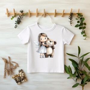 Könnte beinhalten: Weißes T-Shirt mit einer Cartoon-Illustration einer Fee, die einen braunen Teddybären umarmt. Die Fee hat lange braune Haare, ein weißes Kleid und weiße Flügel. Der Teddybär hat einen hellbraunen Körper und ein lächelndes Gesicht. Das Shirt hängt an einem Holzregal.