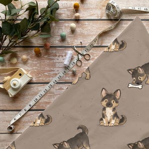 Könnte beinhalten: Rolle Geschenkpapier in Beige mit einem sich wiederholenden Muster von Cartoon-Deutschen Schäferhund-Welpen. Ein Maßband, eine Schere, Faden und eine Spielzeugkamera liegen auf einer Holzoberfläche.