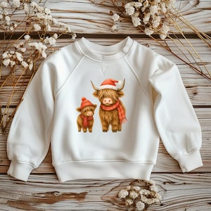 Könnte beinhalten: Weißes Sweatshirt mit einem Aufdruck von zwei Highland-Rindern. Das größere Rind trägt einen Weihnachtsmannhut und einen roten Schal, das kleinere Rind trägt einen passenden Hut und Schal. Die Rinder sind braun mit langem Fell.