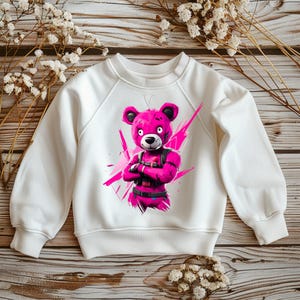 Könnte beinhalten: Weißes Sweatshirt mit einem pinkfarbenen Bären-Motiv, verschränkten Armen und einer schwarzen Weste. Der Bär ist vor einem pinkfarbenen Spritzer-Design platziert. Das Sweatshirt hat lange Ärmel und einen Rundhalsausschnitt.