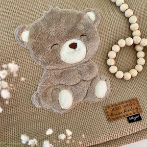 Può includere: Un orsacchiotto marrone con un viso sorridente e dettagli bianchi è appoggiato su una coperta beige strutturata. Sono presenti anche una ghirlanda di perline di legno e piccoli fiori bianchi. Una piccola etichetta marrone recita "Jetzt wird's kuschelig".