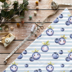 Könnte beinhalten: Blau-weiß gestreifter Stoff mit einem Muster aus lächelnden Blaubeeren. Ein Maßband, eine Schere, Faden und eine Spielzeugkamera liegen auf einer Holzoberfläche. Filzkugeln und Grünzeug sind ebenfalls vorhanden.