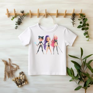 Könnte beinhalten: Weißes T-Shirt mit einer Grafik von drei Cartoon-Charakteren mit unterschiedlichen Haarfarben und Outfits. Die Charaktere halten Waffen oder Werkzeuge. Das T-Shirt hängt an einem Holzgestell mit Grünzeug.