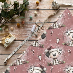 Może przedstawiać: Świąteczny papier do pakowania z powtarzającym się wzorem kreskówkowych szczeniąt w czapkach Świętego Mikołaja. Tło jest w kolorze zakurzonej róży, z laskami cukrowymi i ostrokrzewem. Widoczna jest również miarka, nożyczki i nić.