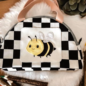 May include: A black and white chequered bag with a pale pink handle. The bag has a white square with an embroidered cartoon bee. The bee is yellow and black with white wings. The bag is on a white fluffy surface.