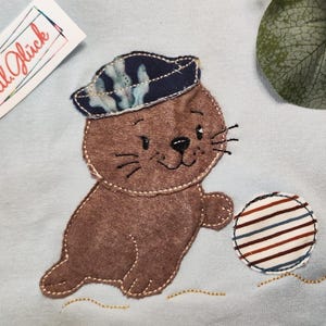 May include: A brown seal applique wearing a blue patterned hat, holding a striped ball. The seal is on a light blue fabric. The image also includes a tag with the text "Textil Glück".