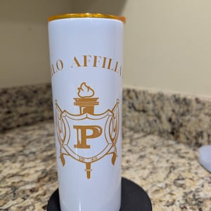 Op de afbeelding: Witte tumbler met gouden accenten en een gouden deksel. De tumbler heeft een gouden embleem met de letter "P" en de woorden "10 Affili" in gouden letters. De tumbler is cilindrisch en staat op een zwarte basis.