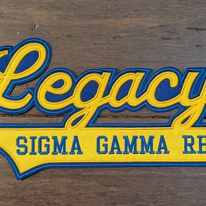 May include: Embroidered patch featuring the word "Legacy" in yellow script with a blue outline. Below, a yellow banner with blue trim reads "SIGMA GAMMA RHO" in blue block letters. The patch is on a wood grain surface.