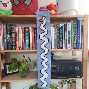 Op de afbeelding: Een blauwe en witte gehaakte wandhanger met een gestileerde zwaan. De hanger wordt getoond op een houten plank met boeken en planten.