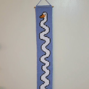 Op de afbeelding: Een gehaakte wandhanger met een witte zwaan met een zwarte omtrek en een oranje snavel. De zwaan is afgebeeld in een golvend, verticaal patroon op een lichtblauwe achtergrond.