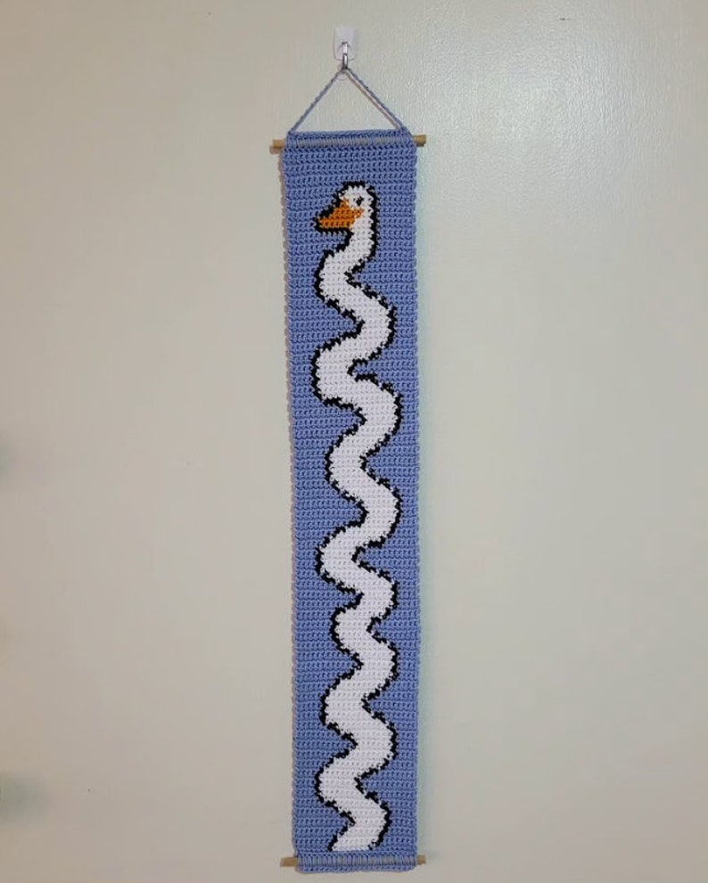 Op de afbeelding: Een gehaakte wandhanger met een witte zwaan met een zwarte omtrek en een oranje snavel. De zwaan is afgebeeld in een golvend, verticaal patroon op een lichtblauwe achtergrond.