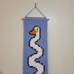 Op de afbeelding: Een gehaakte wandhanger met een witte gans met een oranje snavel op een lichtblauwe achtergrond. De hanger is bevestigd aan een houten stok.