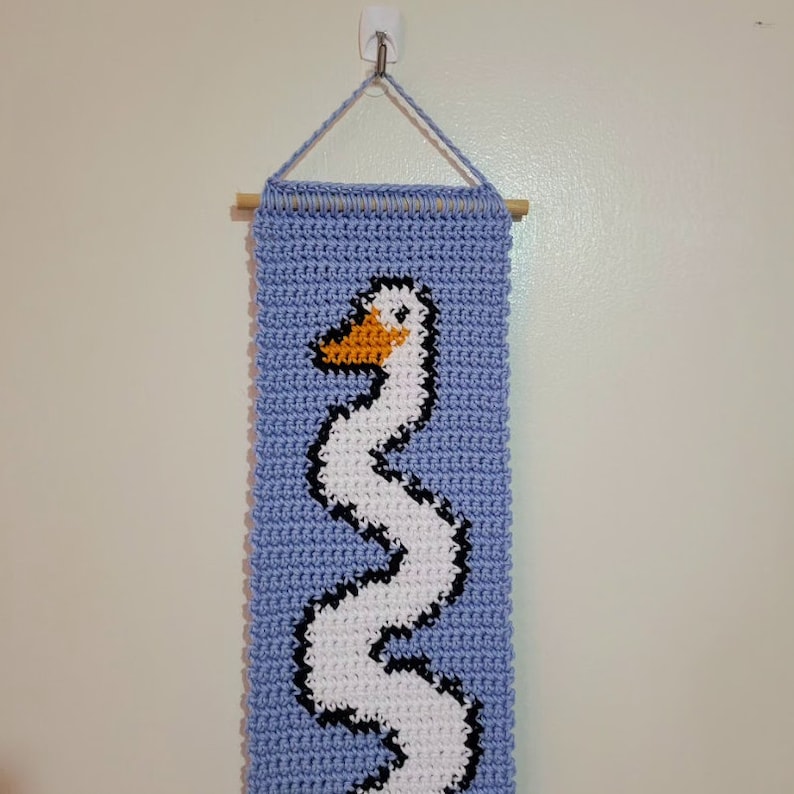 Op de afbeelding: Een gehaakte wandhanger met een witte gans met een oranje snavel op een lichtblauwe achtergrond. De hanger is bevestigd aan een houten stok.