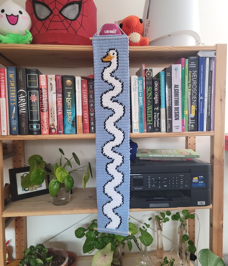 Op de afbeelding: Een blauwe en witte gehaakte wandhanger met een gestileerde zwaan. De hanger wordt getoond op een houten plank met boeken en planten.