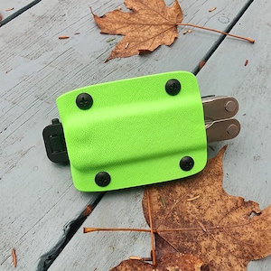 May include: A bright green multi-tool holster with black snaps and a black clip. The holster is lying on a wooden surface with brown leaves.