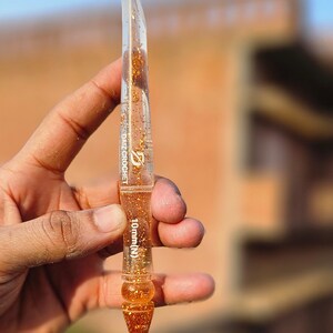 May include: A clear resin crochet hook featuring gold glitter flakes embedded throughout the body. The tool displays the text DAIZ CROCHET and 10mm (N) printed in white on the shaft. The hook has a tapered point and a decorative pointed end.