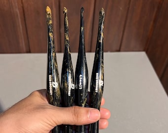 Puede incluir: Cuatro ganchillos con un acabado jaspeado en negro y dorado. Cada ganchillo presenta un mango cónico y una punta. El texto "DAIZ CROCHET" está impreso en tres de los mangos, mientras que uno muestra el texto "4.5mm(7)" en letras blancas.