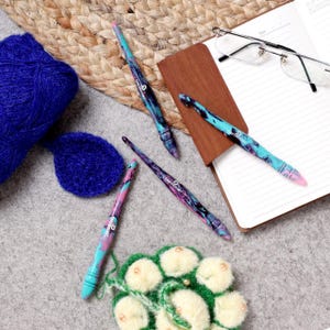 Puede incluir: Cuatro ganchillos coloridos con un patr&oacute;n de remolino, dispuestos sobre una superficie gris con un cuaderno, gafas y lana.
