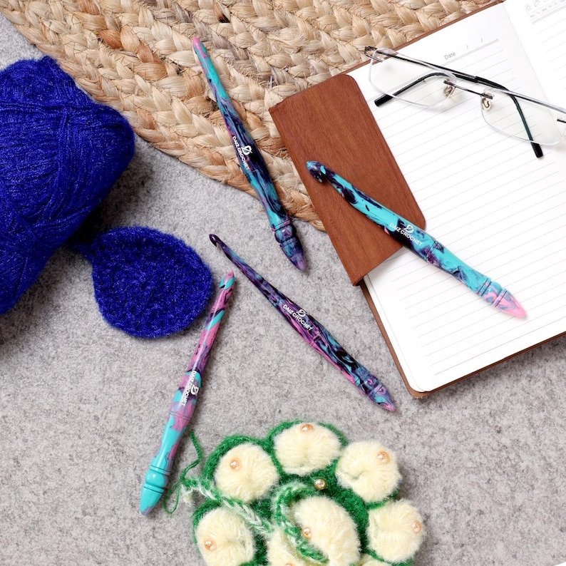 Puede incluir: Cuatro ganchillos coloridos con un patr&oacute;n de remolino, dispuestos sobre una superficie gris con un cuaderno, gafas y lana.