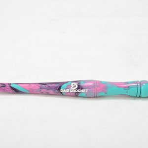 Puede incluir: Un gancho de ganchillo de color turquesa, rosa y morado con un logotipo "DAIZ CROCHET" en negro.