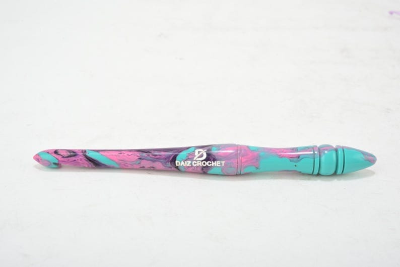 Puede incluir: Un gancho de ganchillo de color turquesa, rosa y morado con un logotipo "DAIZ CROCHET" en negro.