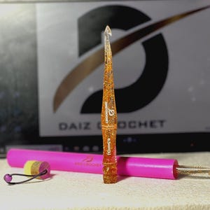 May include: A clear resin crochet hook with orange and gold glitter suspended inside, featuring the text DAIZ CROCHET and 6.5mm(K) printed on the shaft. It is positioned vertically next to a bright pink cylindrical case with a small yellow stopper.