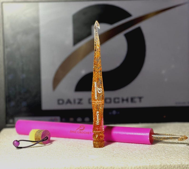 May include: A clear resin crochet hook with orange and gold glitter suspended inside, featuring the text DAIZ CROCHET and 6.5mm(K) printed on the shaft. It is positioned vertically next to a bright pink cylindrical case with a small yellow stopper.