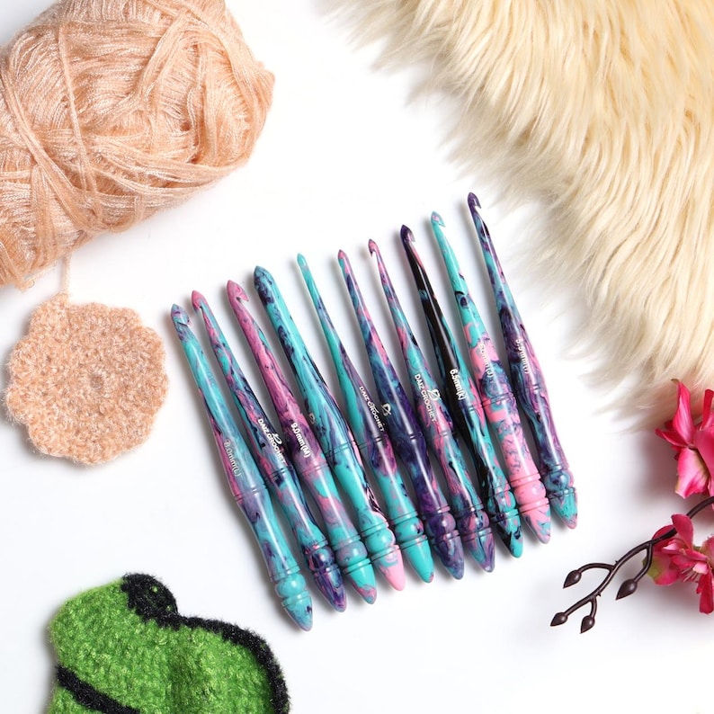 Puede incluir: Conjunto de diez ganchillos de crochet coloridos con un dise&ntilde;o de m&aacute;rmol en tonos de rosa, azul y morado. Los ganchillos son de madera y tienen un acabado liso.