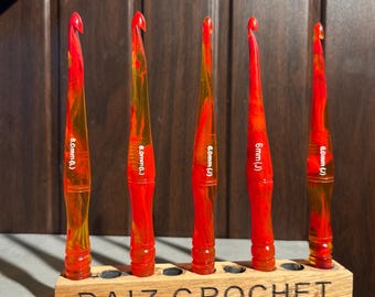 Puede incluir: Un juego de seis ganchillos de acrílico jaspeado rojo y amarillo. Cinco ganchillos están colocados verticalmente en un bloque de madera con la etiqueta DAIZ CROCHET, con las tallas 8.0mm (L) y 6.0mm (J). Uno yace horizontalmente delante.