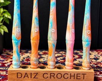 May include: Six crochet hooks with a marbled pattern of blue, orange, and pink, each featuring the text DAIZ CROCHET. Five hooks stand upright in a rectangular wooden holder engraved with DAIZ CROCHET, while one hook lies horizontally in front.