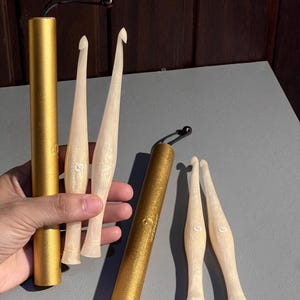 May include: Four cream-colored crochet hooks with a pearlescent finish and two cylindrical gold storage cases. Each hook features a tapered handle and a small white logo printed on the shaft. The cases have black cords attached to the top caps.