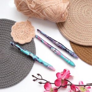 Puede incluir: Tres ganchillos de crochet coloridos con un dise&ntilde;o de m&aacute;rmol y el texto "Bazi Crochet" en cada gancho. Los ganchillos est&aacute;n sobre una superficie blanca con una flor rosa, una bola de hilo y dos c&iacute;rculos de crochet.