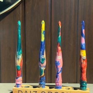 Könnte beinhalten: Vier mehrfarbige Häkelnadeln aus Kunstharz mit konischen Griffen, die jeweils den Schriftzug DAIZ CROCHET und ein Logo tragen. Die Nadeln stehen aufrecht in einem rechteckigen Holzständer, in dessen Vorderseite DAIZ CROCHET eingraviert ist.