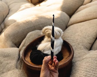 May include: A black and white swirl patterned crochet hook with the text Furls Crochet printed on the side. The hook is held vertically in front of a wooden bowl containing a ball of white yarn and a ball of black yarn.