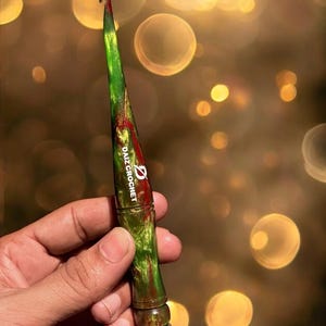 Puede incluir: Un ganchillo verde y rojo con el texto "DAIZ CROCHET" impreso en el lateral. El ganchillo se sostiene en una mano sobre un fondo borroso de luces bokeh doradas. El ganchillo mide aproximadamente 15 cm de largo.